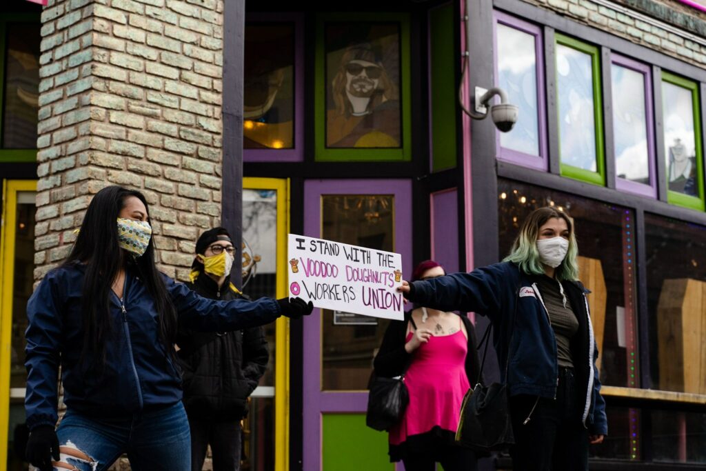 Dos trabajadoras llevan una pancarta que dice "Estoy con el Sindicato de Trabajadores de Voodoo Donut".