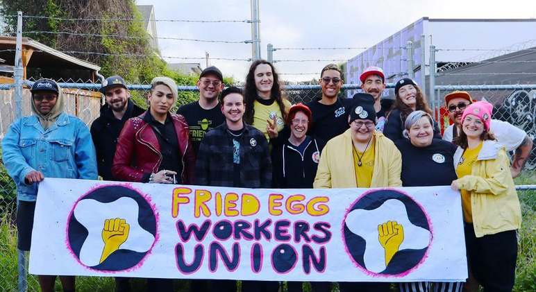 Catorce trabajadores alegres de Fried Egg I’m in Love en Portland, Oregon, Estados Unidos, con su banner que dice, Fried Egg Workers Union.