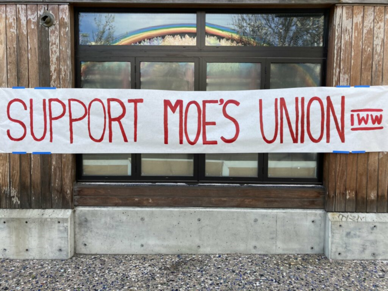 Foto por Zack Huber mostrando una pancarta sobre una ventana que refleja un arcoíris. La pancarta dice, Apoye al Sindicato de Moe's, TIM.
