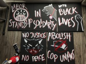 Letreros de piquete en ingles que dicen, Sindicatos se levantan en solidaridad con las Vida Negras. Otro letrero dice No justicia, no paz. Otro dice Abolir los sindicatos de policía. 