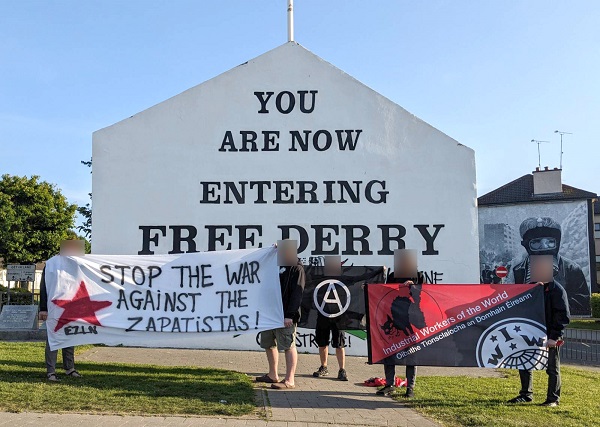 Miembros de los TIM en Derry manifestando su solidaridad con las comunidades Zapatistas en Chiapas, México. Están frente de una casa con un mural que dice Ya esta entrando a Derry Libre.